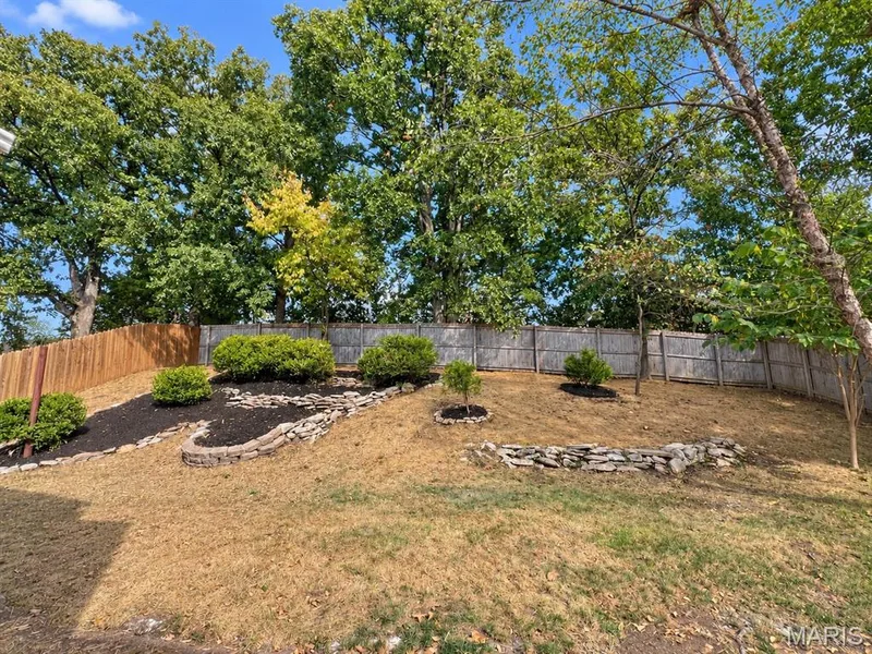 Backyard featuring a wood privacy fence, mature trees, landscaped planting beds with mulch, and stone retaining walls — 1731 Fairview Farms Circle, Wentzville, MO 63385