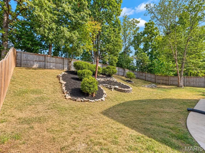 Expansive backyard with wood fencing, lush trees, stone-edged landscaping beds, and a concrete patio section — 1731 Fairview Farms Circle, Wentzville, MO 63385
