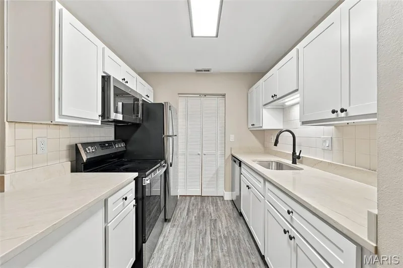 Galley kitchen featuring white cabinetry with black hardware, light-toned countertops, tile backsplash, and wood-finish flooring — 2415 Sandalwood Creek Court #C, Wildwood, MO 63038