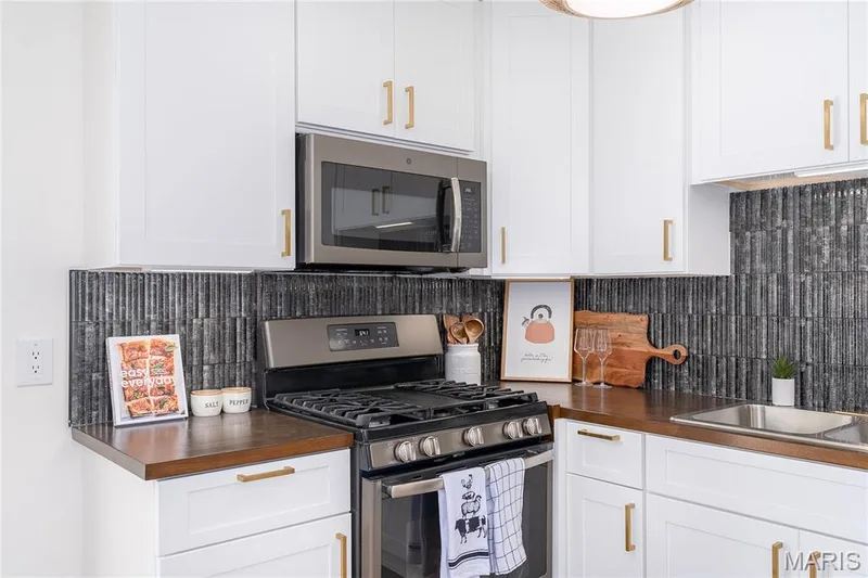 Stainless steel gas range with overhead microwave, white cabinetry with gold-tone hardware, dark wood-finish countertops, and a dark tile backsplash — 3025 Sherwood Lane, St Charles, MO 63301