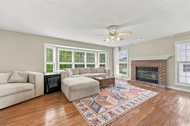 Hardwood flooring, bay window with multiple panes, brick fireplace with mantel, ceiling fan with light fixture, and multiple windows with blinds — 8 Jacobs Place Court, O'Fallon, MO 63376