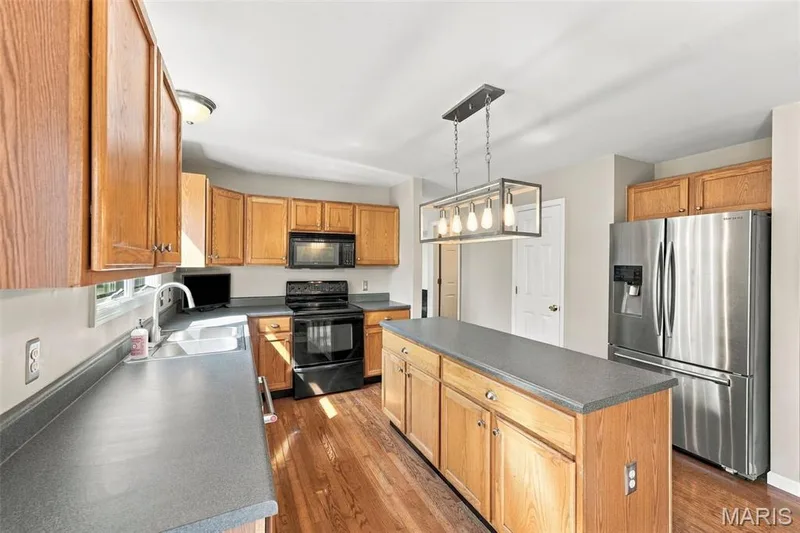 Kitchen featuring wood-finish cabinetry, stainless steel appliances, a center island with countertop space, wood-finish flooring, and a modern rectangular pendant light fixture — 8 Jacobs Place Court, O'Fallon, MO 63376