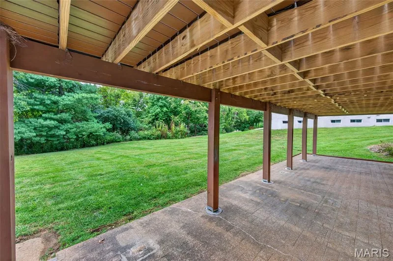 Covered outdoor patio with a stamped concrete surface, supported by substantial posts, overlooking a expansive grassy area bordered by mature trees. — 231 Renaldo Drive, Chesterfield, MO 63017