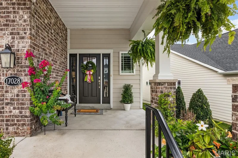 Welcoming front porch featuring a dark-finish entry door with sidelights, brick exterior with climbing magenta flowers, hanging fern planters, potted topiaries, and a concrete landing with black metal railings — 17028 Fire Glow Drive, Ballwin, MO 63011