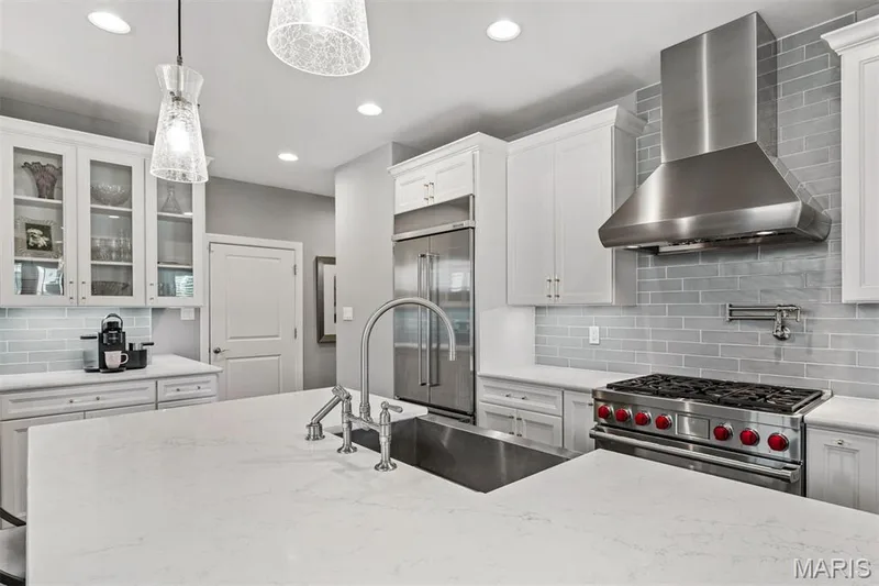 Chef's kitchen featuring a professional-grade gas range with red knobs, stainless steel range hood, undermount sink with modern faucet, and white cabinetry with glass-front upper units — 17028 Fire Glow Drive, Ballwin, MO 63011