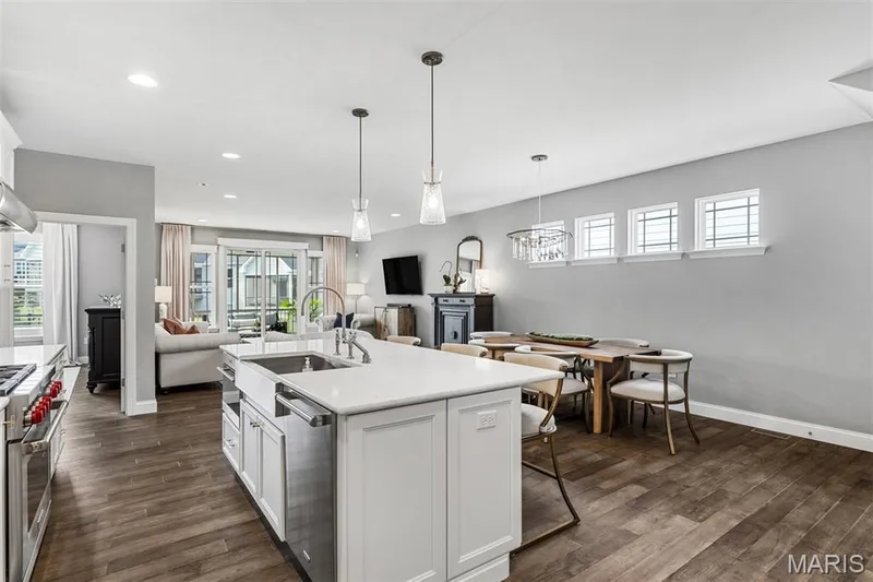 Open-concept kitchen and dining area featuring a large white cabinetry island with waterfall countertop, stainless steel appliances, wood-finish flooring throughout, recessed and pendant lighting, and abundant natural light from transom windows — 17028 Fire Glow Drive, Ballwin, MO 63011