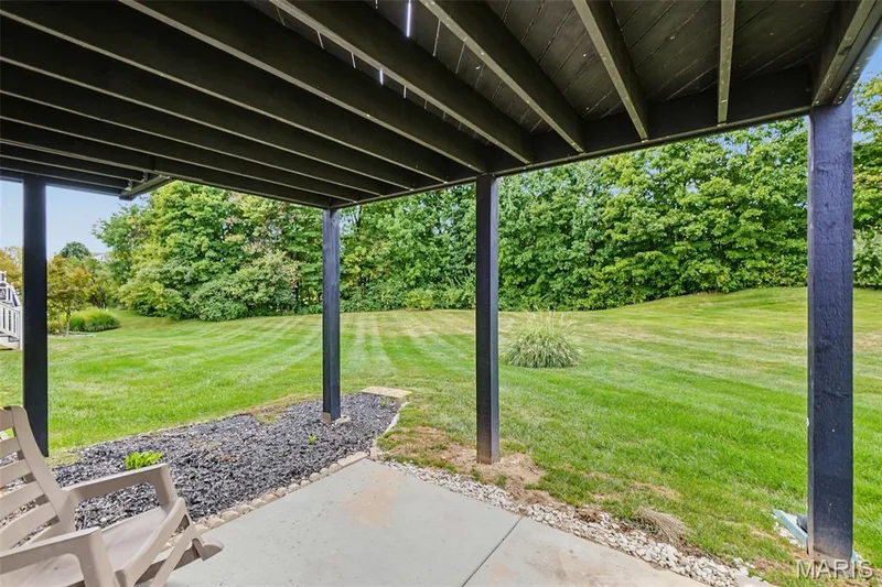 Covered patio area with concrete surfacing, black wood support beams, and landscaping featuring dark stone and a manicured lawn — 8 Soenker Circle, St Peters, MO 63376