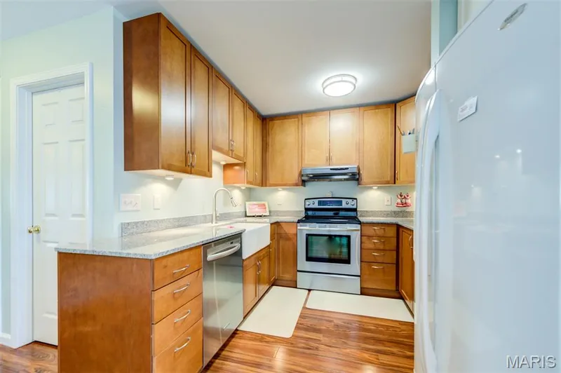 Kitchen featuring wood-finish flooring, light wood cabinetry, white farmhouse sink, stainless steel appliances, and granite countertops — 820 Brookmead Drive, O'Fallon, MO 63366