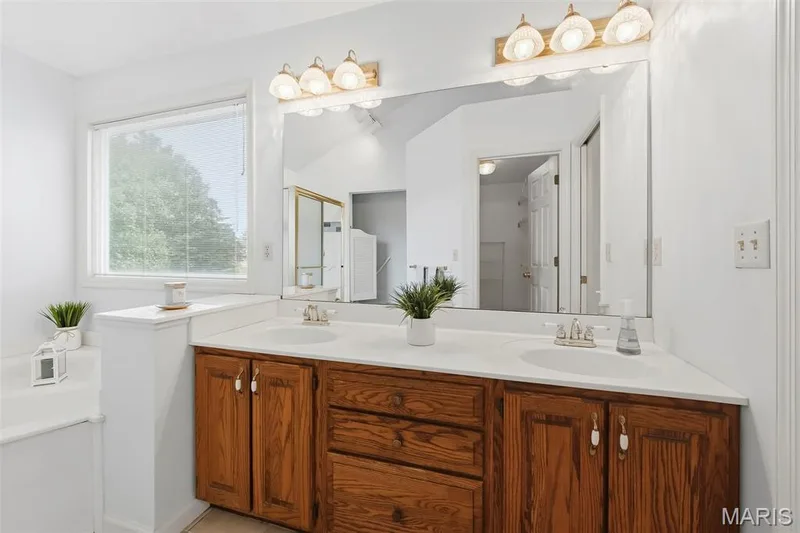 Dual vanity with integrated sinks, wood-finish cabinetry, a large mirror, and overhead lighting — 11 Silver Leaf Court, Lake St. Louis, MO 63367