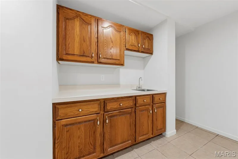 Wood-finish cabinetry with brass hardware, light-toned countertop, stainless steel sink with chrome faucet, and neutral tile flooring — 11 Silver Leaf Court, Lake St. Louis, MO 63367
