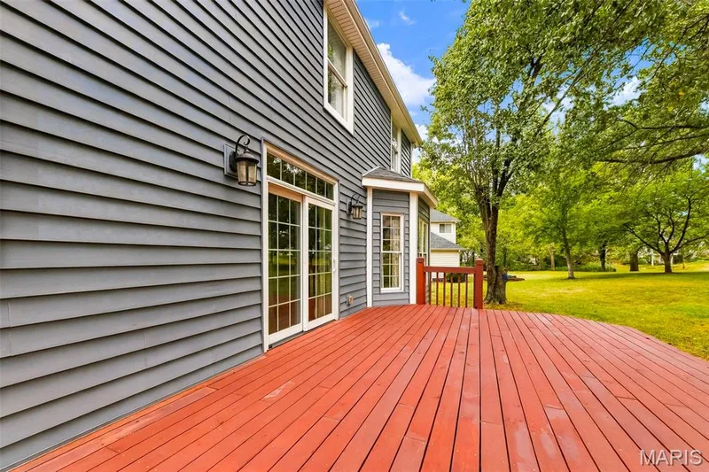 Expansive wood deck with red finish, gray horizontal siding, exterior wall sconces, sliding glass patio doors, and a partial bay window — 11 Silver Leaf Court, Lake St. Louis, MO 63367
