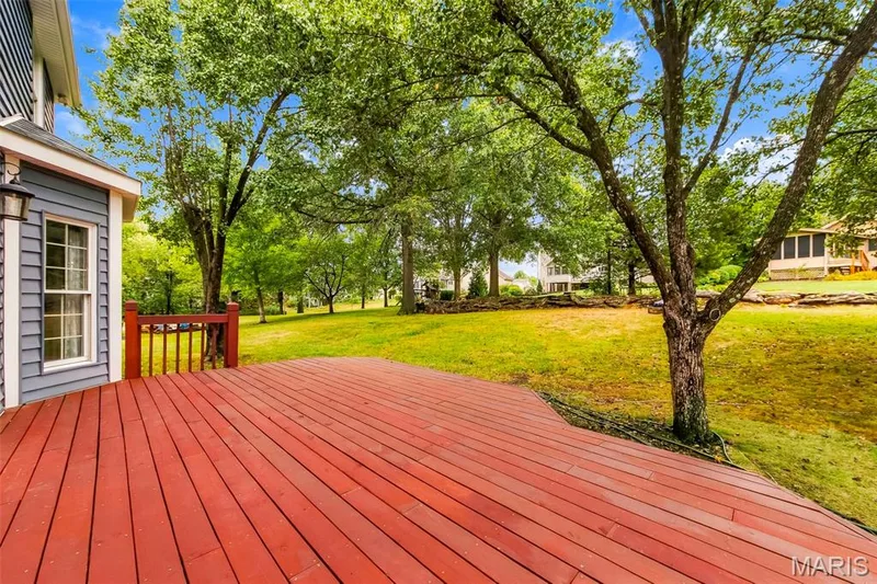 Expansive wood deck with a red finish, featuring a coordinating wood railing and a built-in exterior light fixture, overlooking a large green lawn with mature trees — 11 Silver Leaf Court, Lake St. Louis, MO 63367