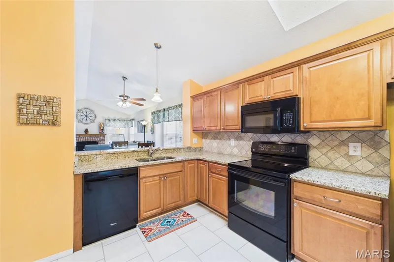 Kitchen featuring light wood cabinetry, granite countertops, integrated sink, black appliances, and a travestine tiled backsplash — 2931 Plum Leaf Circle, St Peters, MO 63303