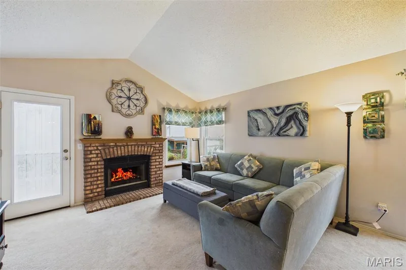 Vaulted ceiling, brick fireplace with a wood mantle, carpeted flooring, and a glass-paneled door exiting to the back deck. — 2931 Plum Leaf Circle, St Peters, MO 63303