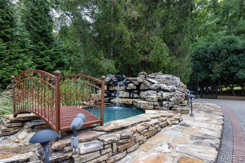 Stone-lined water feature with a waterfall, decorative metal bridge, and natural stone surround — 76 River Valley Drive, Chesterfield, MO 63017