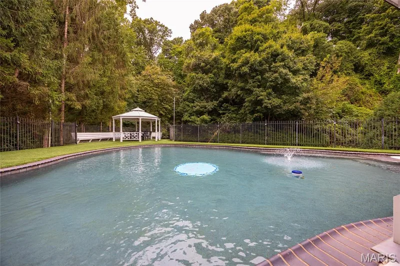 Curved swimming pool with an integrated fountain, surrounded by a black metal fence, and lush mature trees — 76 River Valley Drive, Chesterfield, MO 63017