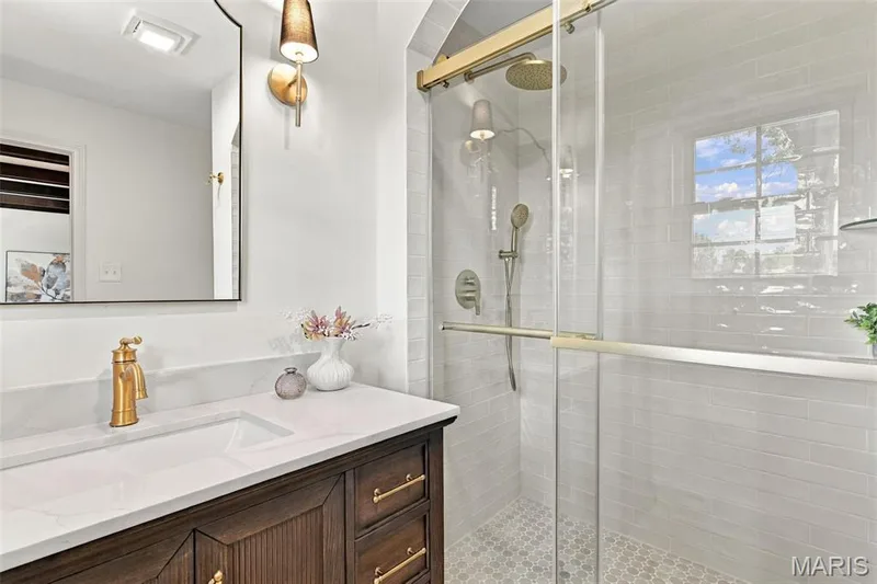 Built-in vanity with a white countertop and undermount sink, gold-tone faucet, framed mirror, and enclosed shower with a gold-tone rainfall showerhead — 621 Claymont Estates Drive, Chesterfield, MO 63017
