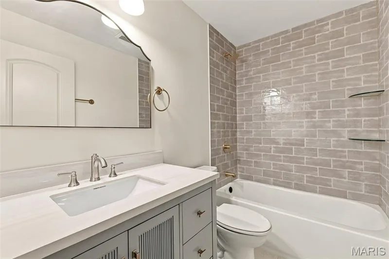 White solid surface countertop with an integrated sink, gray shaker-style vanity with reeded paneling, an arched mirror, a white bathtub, and subway tile shower surround with glass shelving — 621 Claymont Estates Drive, Chesterfield, MO 63017