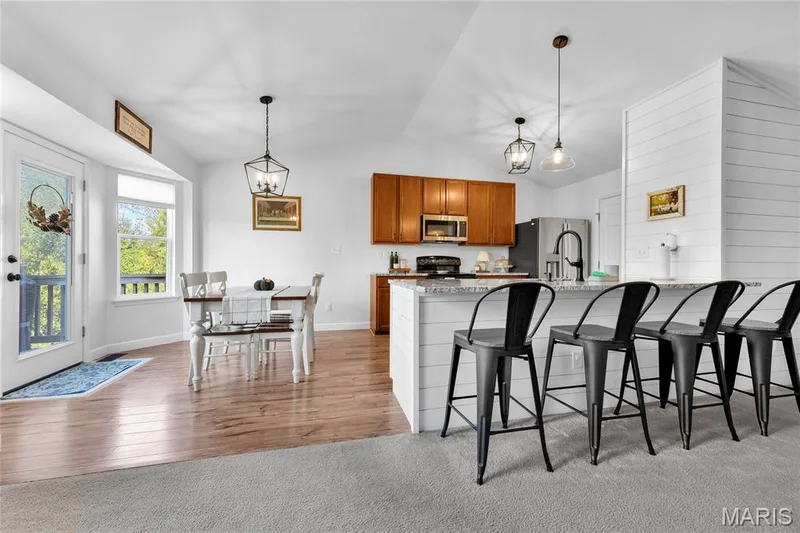 Vaulted ceiling with decorative lighting fixtures, wood-finish flooring, kitchen island with shiplap-style siding, light-toned countertops, and wood cabinetry — 5507 Dressel Drive, Cottleville, MO 63304