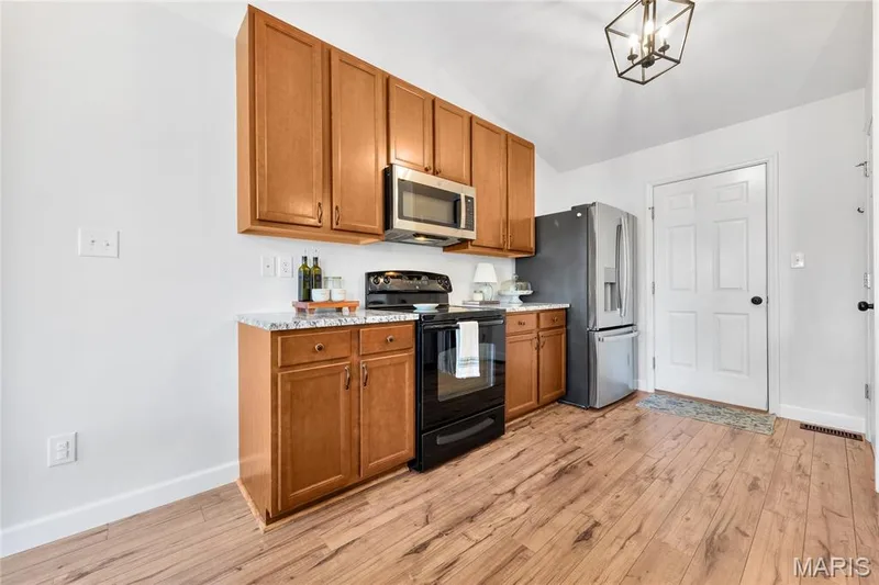 Wood-finish flooring, wood cabinetry with brushed nickel hardware, stainless steel appliances, granite-style countertops, and a vaulted ceiling with a decorative light fixture — 5507 Dressel Drive, Cottleville, MO 63304