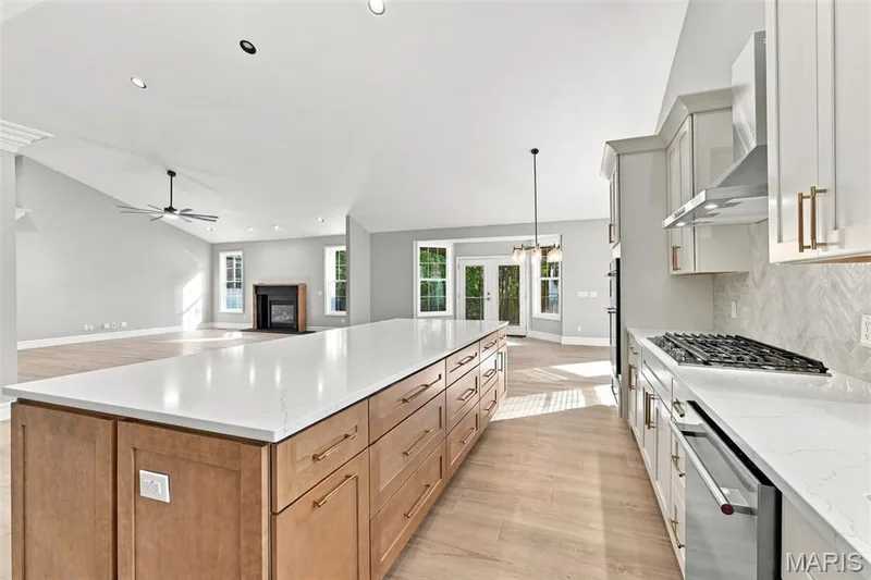 Spacious kitchen featuring a large central island with light wood-finish cabinetry, a gas range, stainless steel Kitchen Aid appliances, and a herringbone-patterned backsplash — 1580 Madre Drive, Foristell, MO 63348