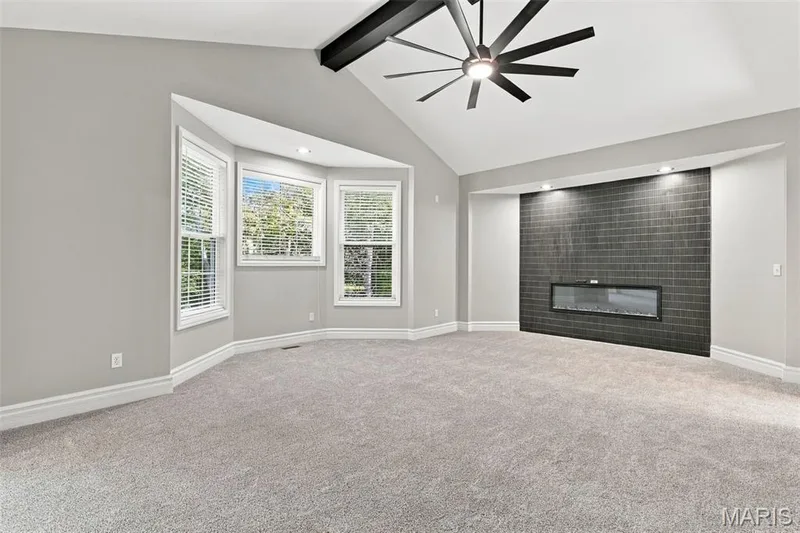 Vaulted ceiling with an exposed beam, contemporary ceiling fan, bay window with blinds, inset electric fireplace with a tiled surround, and light-toned carpeting — 1580 Madre Drive, Foristell, MO 63348