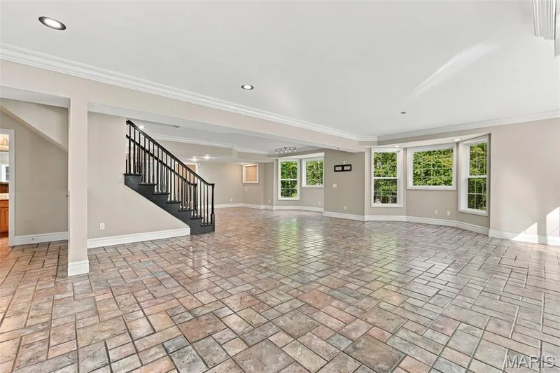Spacious lower level featuring patterned tile flooring, a black staircase with wood banister, multiple windows with a treed backdrop, and recessed lighting — 1580 Madre Drive, Foristell, MO 63348