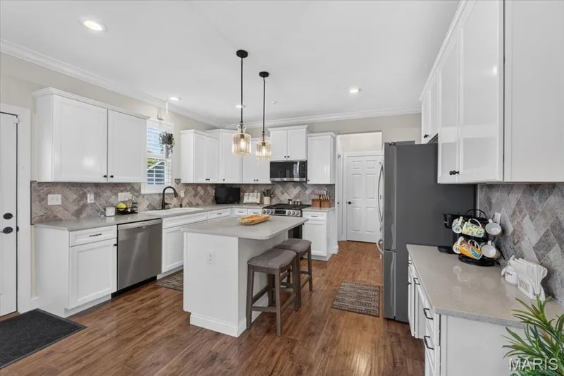 Kitchen featuring wood-finish flooring, white cabinetry, a central island with seating, stainless steel appliances, and a herringbone pattern tile backsplash — 3708 Broad Street, St Charles, MO 63301