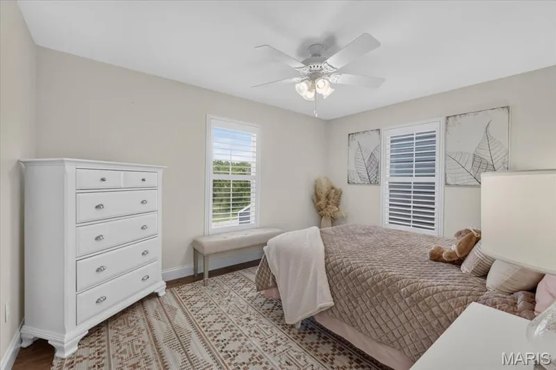 Warm neutral paint throughout, ceiling fan with integrated lighting, wood-finish flooring, two large windows with louvered shutters, and a streamlined white dresser with metal hardware — 3708 Broad Street, St Charles, MO 63301