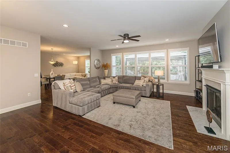 Warm wood-finish flooring, recessed lighting, ceiling fan, fireplace with white mantel, and multiple windows with white louvered blinds — 1229 Cashmere Lane, St Peters, MO 63376