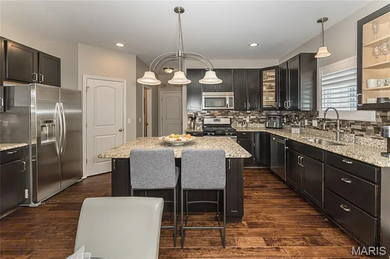 Expansive kitchen featuring dark wood cabinetry, stainless steel appliances, a central island with countertop seating, and wood-finish flooring — 1229 Cashmere Lane, St Peters, MO 63376