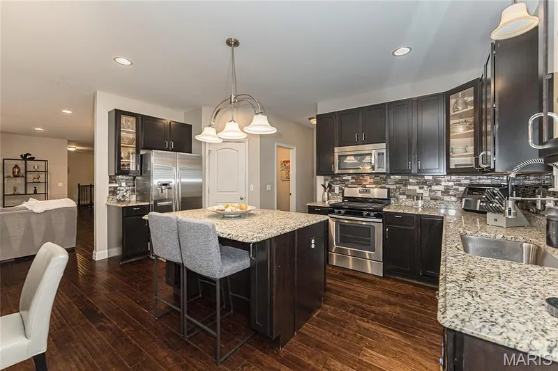 Kitchen featuring dark wood-finish cabinetry, granite countertops, stainless steel appliances, wood-finish flooring, and a mosaic tile backsplash — 1229 Cashmere Lane, St Peters, MO 63376