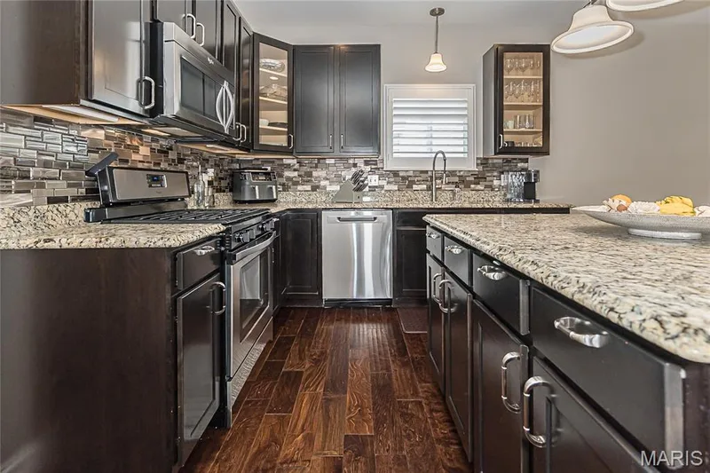 Kitchen featuring dark wood cabinetry, light-toned granite countertops, a mosaic tile backsplash, stainless steel appliances, and wood-finish flooring — 1229 Cashmere Lane, St Peters, MO 63376