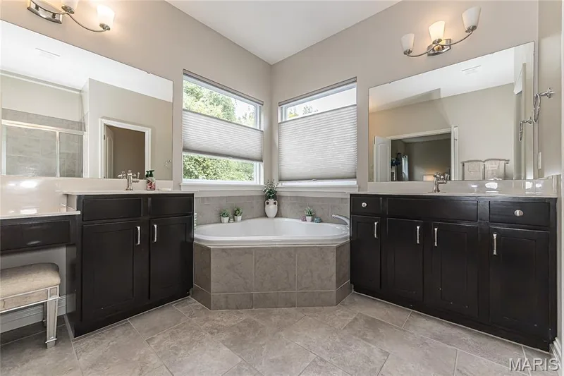Dual dark wood-finish vanities with light-toned countertops, corner soaking tub with tiled surround, and large wall-mounted mirrors — 1229 Cashmere Lane, St Peters, MO 63376