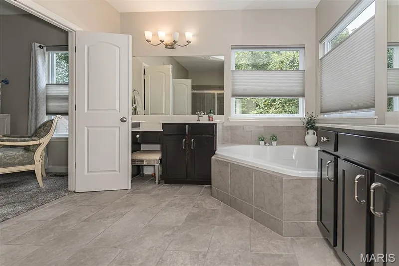Spacious bathroom featuring a built-in corner soaking tub, dual dark wood vanities, tiled flooring, and multiple windows — 1229 Cashmere Lane, St Peters, MO 63376