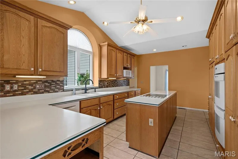Wood-finish cabinetry with white countertops, tile flooring, built-in double ovens, and a vaulted ceiling with a ceiling fan — 104 Wolf Trail Court, Defiance, MO 63341