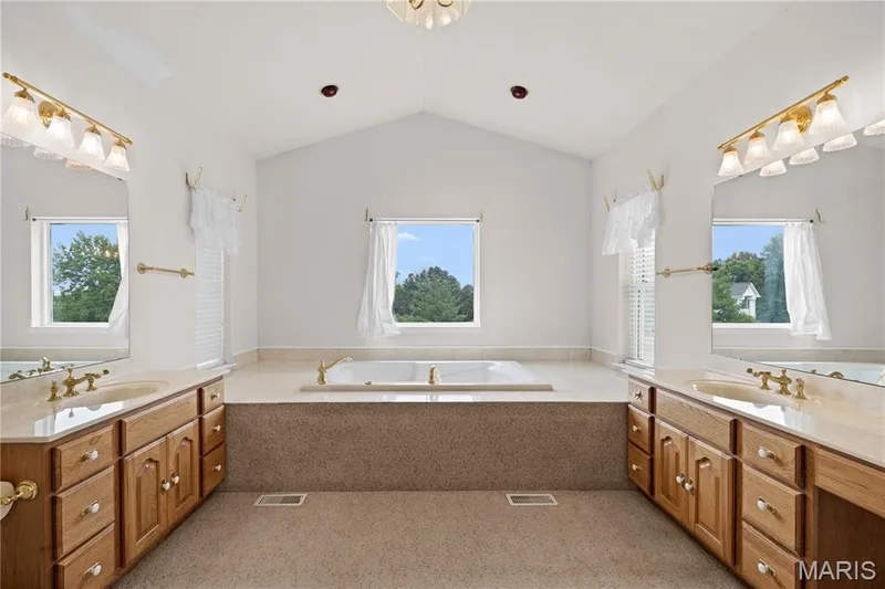 Vaulted ceiling bathroom featuring a built-in tub, dual vanity with wood cabinetry, and integrated windows — 104 Wolf Trail Court, Defiance, MO 63341
