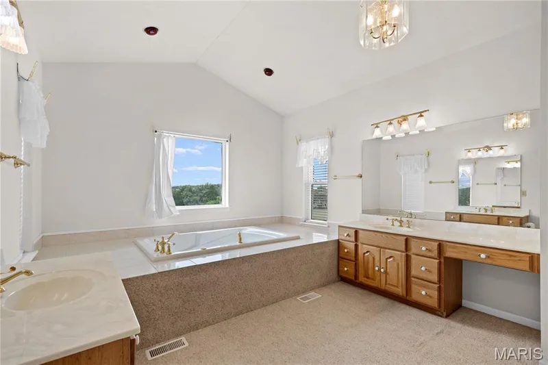 Vaulted ceiling, built-in soaking tub, dual vanities with integrated sinks, wood cabinetry, and a large wall-mounted mirror — 104 Wolf Trail Court, Defiance, MO 63341