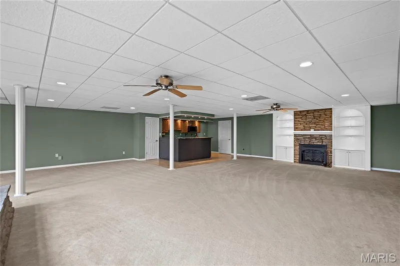 Expansive carpeted area featuring a stone-faced fireplace with built-in shelving, a recessed wet bar, and a drop ceiling with recessed lighting and ceiling fans — 104 Wolf Trail Court, Defiance, MO 63341