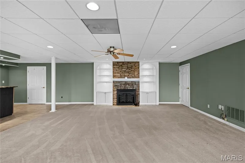 Expansive carpeted area featuring a stone-clad fireplace with mantel, built-in arched shelving, an integrated ceiling fan, and recessed lighting — 104 Wolf Trail Court, Defiance, MO 63341