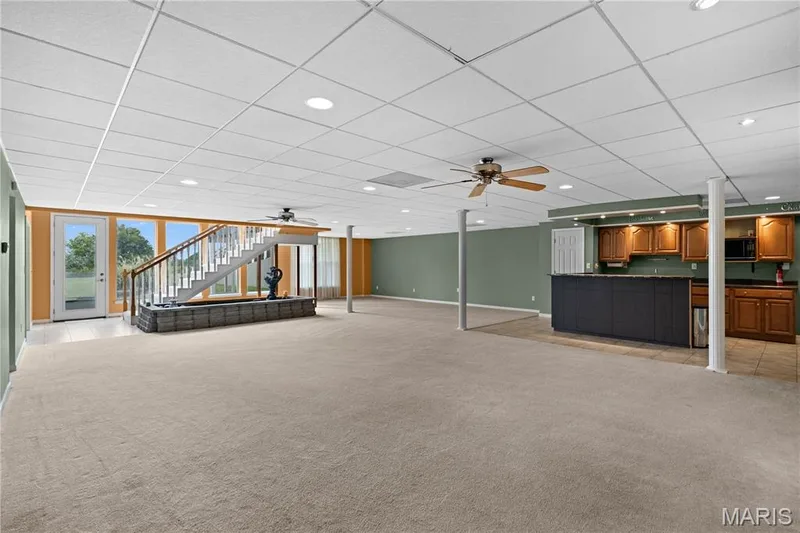 Expansive open-concept room with carpet flooring, a built-in wet bar featuring wood cabinetry and dark countertops, recessed lighting, and a white open staircase with wood handrails — 104 Wolf Trail Court, Defiance, MO 63341