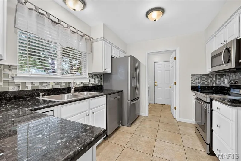 Kitchen featuring white cabinetry, dark speckled countertops, a mosaic tile backsplash, stainless steel appliances, and tiled flooring — 20 Autumn Ridge Drive #83B, O'Fallon, MO 63366