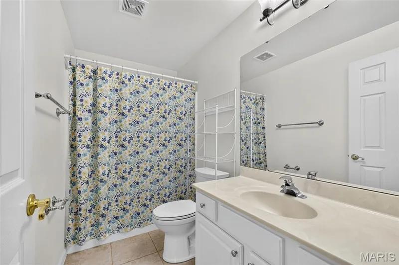 Vanity with an integrated sink and chrome faucet, large wall-mounted mirror, white paneled door, and tiled flooring — 20 Autumn Ridge Drive #83B, O'Fallon, MO 63366