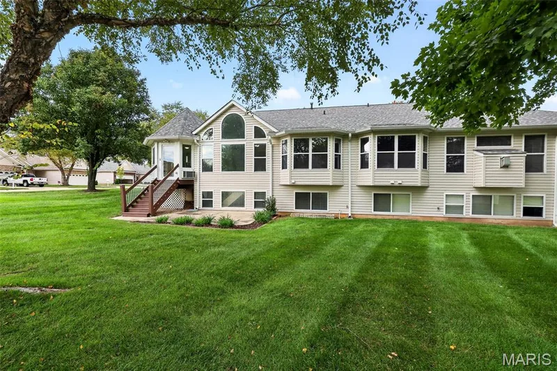 Expansive green lawn with mature trees, a multi-level deck with wooden railings, and a stone patio — 20 Autumn Ridge Drive #83B, O'Fallon, MO 63366