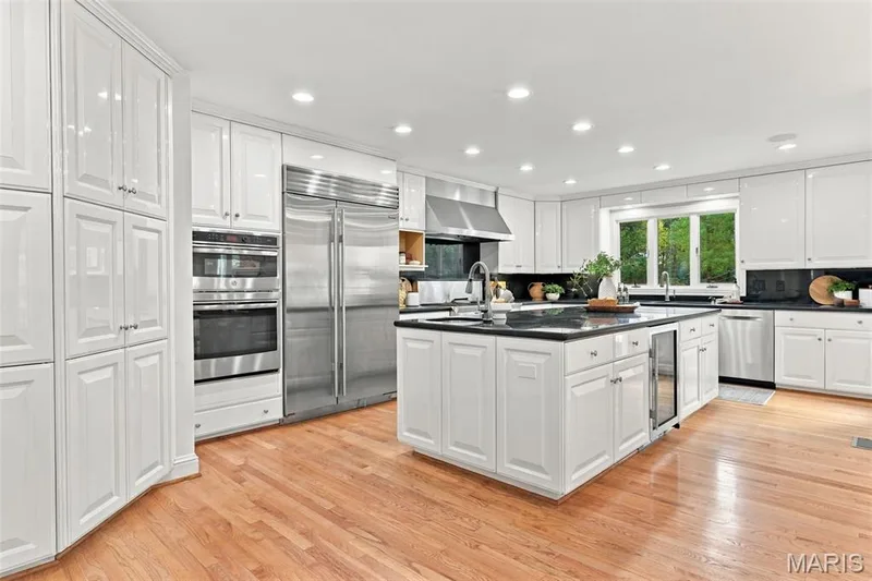 White cabinetry with polished hardware, stainless steel appliances, central island with an under-counter beverage cooler, dark countertops, and wood-finish flooring — 2785 Sun Meadow Drive, Wildwood, MO 63005