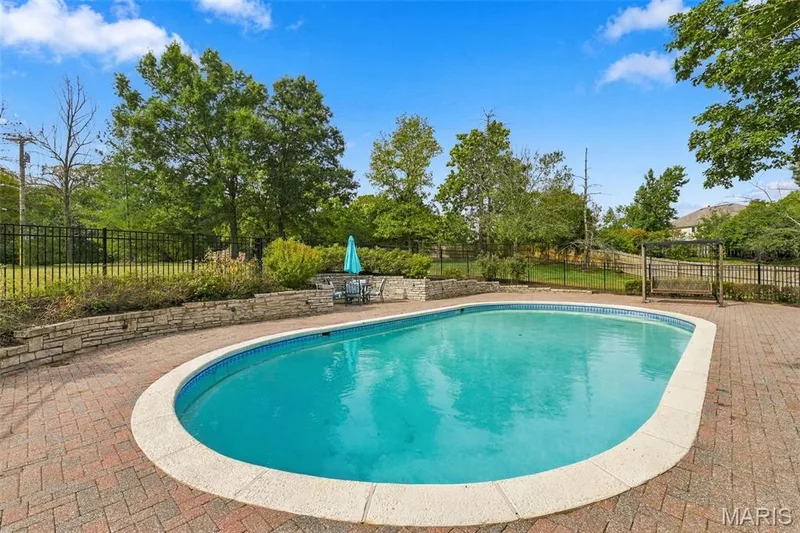 Oval-shaped swimming pool with a brick paver surround, stone retaining walls, and a black wrought iron fence — 2785 Sun Meadow Drive, Wildwood, MO 63005