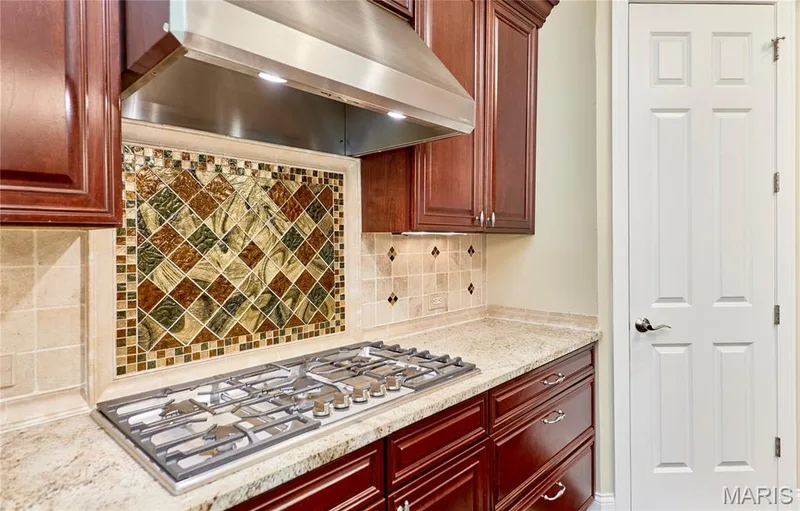 Stainless steel range hood with integrated lighting, a gas cooktop, rich wood-finish cabinetry, and a decorative tile backsplash with Murano glass mosaic detailing — 1970 Chesterfield Ridge Circle, Chesterfield, MO 63017