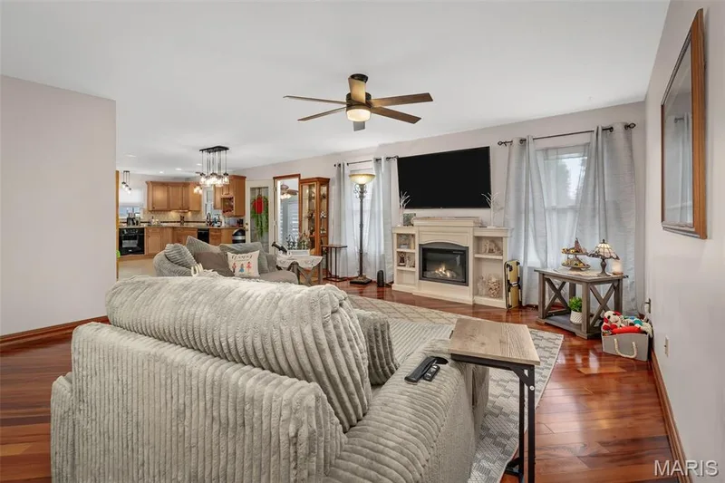 Hardwood flooring, ceiling fan, fireplace with built-in shelving, recessed lighting, and a kitchen visible in the background — 355 Birmingham Drive, O'Fallon, MO 63366
