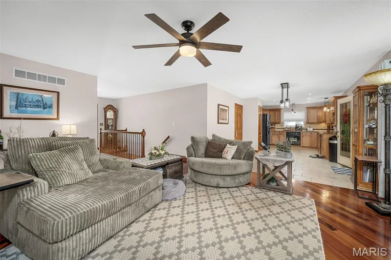 Open concept living area featuring wood-finish flooring, a ceiling fan with integrated lighting, and a partial staircase with wood railing — 355 Birmingham Drive, O'Fallon, MO 63366