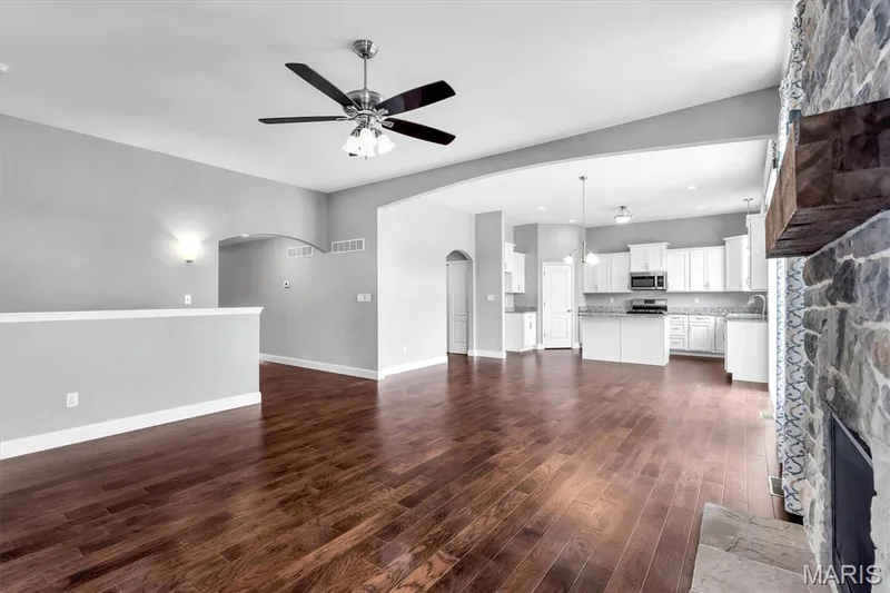 Wood-finish flooring throughout, vaulted ceiling with a ceiling fan, stone-faced fireplace with a wood mantle, white kitchen cabinetry, and pendant lighting — 332 Mystic Brook Drive, O'Fallon, MO 63366